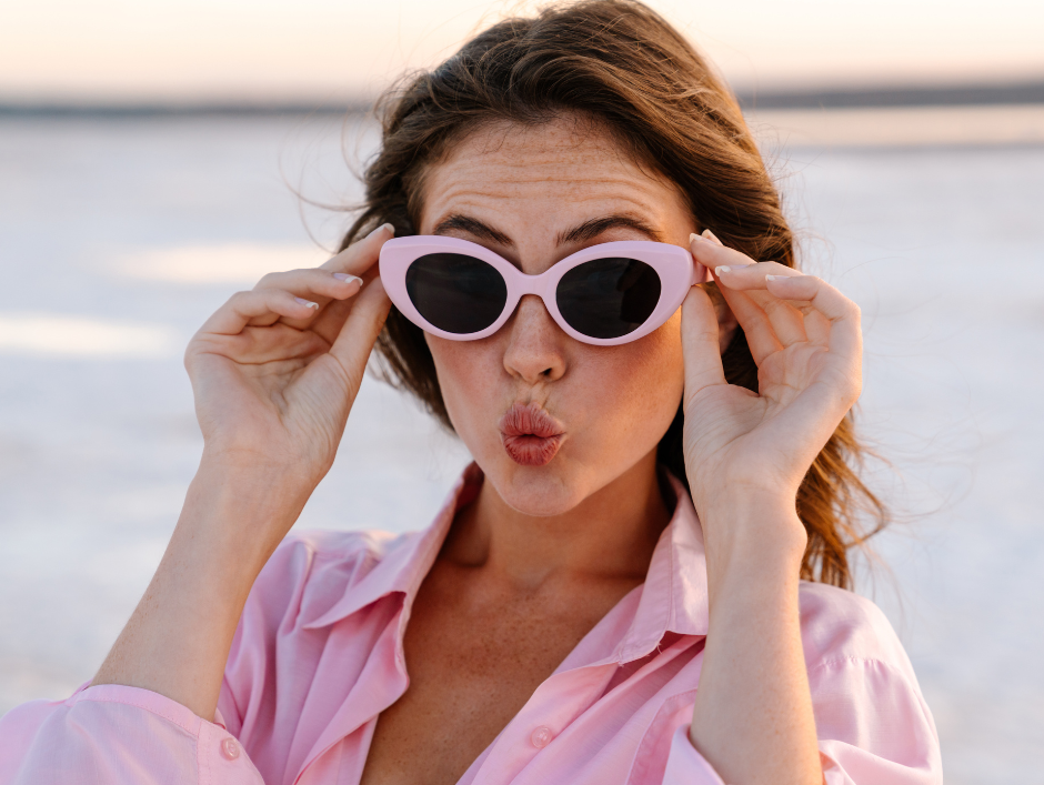 A lady standing in front of the sea is wearing a pink shirt and pink sunglasses