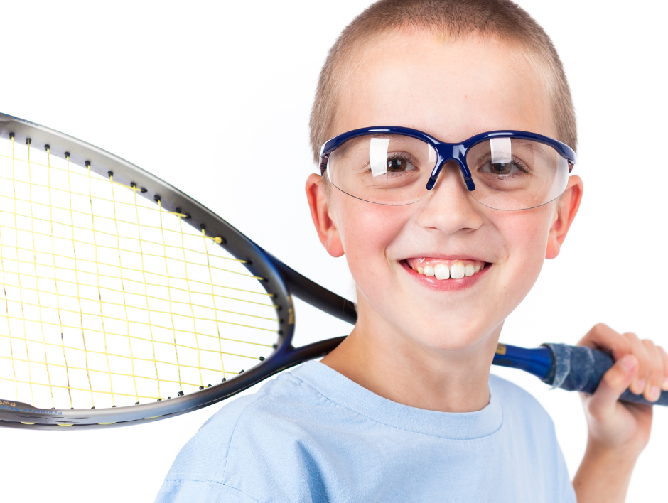 A boy wearing protective eye glasses, he is holding a tennis racquet over his shoulder..