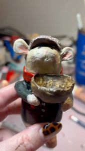 A handmade white mouse. The mouse is wearing a flat cap, coat and red scarf and is holding an empty bowl with outstreached arms.