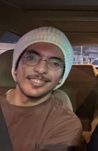 Photo of Saud who is sitting in a car, smiling at the camera. They are wearing a white beanie, glasses, and a brown top. The background shows the interior of the car with faint details of a street visible through the windows.