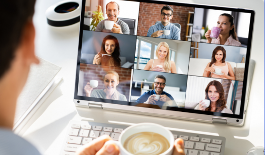 A person is facing a laptop holding a mug of coffee, on the screen are nine people all facing the camera.