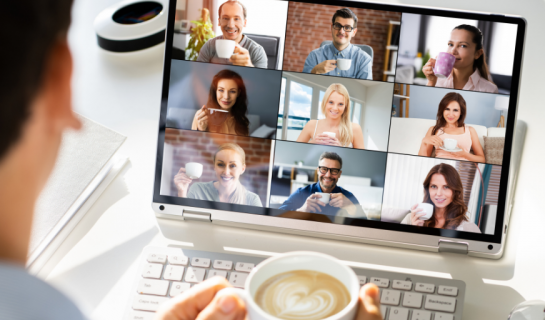 A person is facing a laptop holding a mug of coffee, on the screen are nine people all facing the camera.