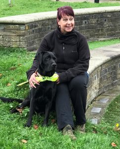 Tanvir is sat outside on a low wall. She is smiling and looking to the right. Her black guide dog is sat in front of her.