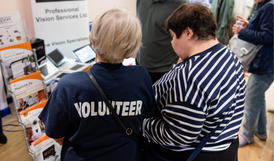 A photo from one of our events. From the back: a lady wearing on of our Volunteer tshirts is assisting a lady by guiding and explaining the equipment in front of them.